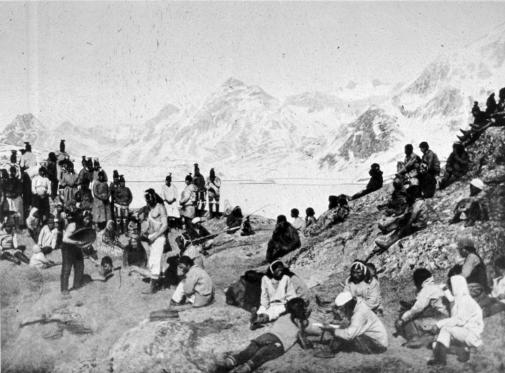 A Greenlandic Inuit community gathered on the rocky shore of a fjord, snow mountains behind