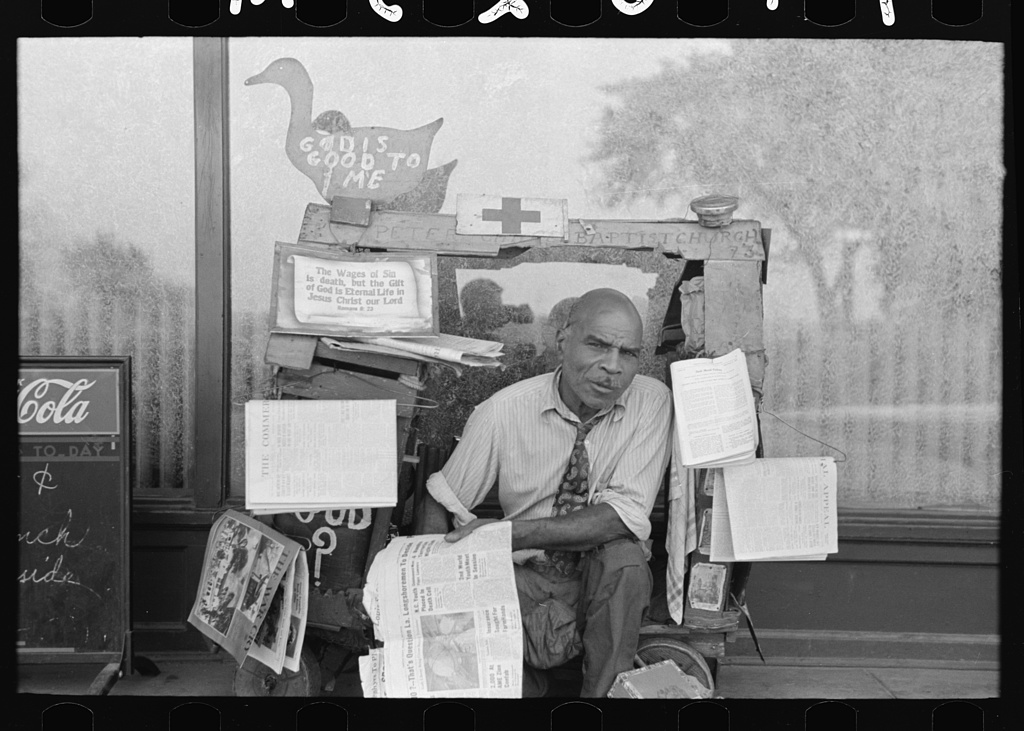 A street vendor surrounded by stacks of newspapers and hand-lettered signs