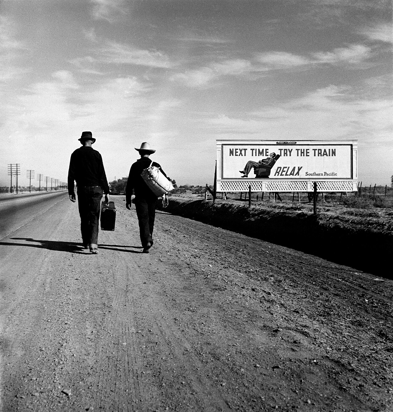Two men walk away down an empty highway past a billboard reading "Next time try the train"
