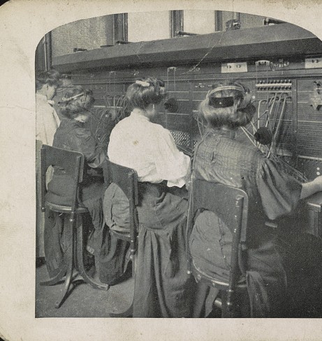 Telephone switchboard operators seen from behind, working a wall of jacks and cords