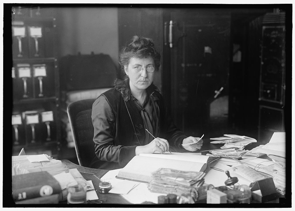 A clerk at a desk piled with papers and ledgers, files and a safe behind