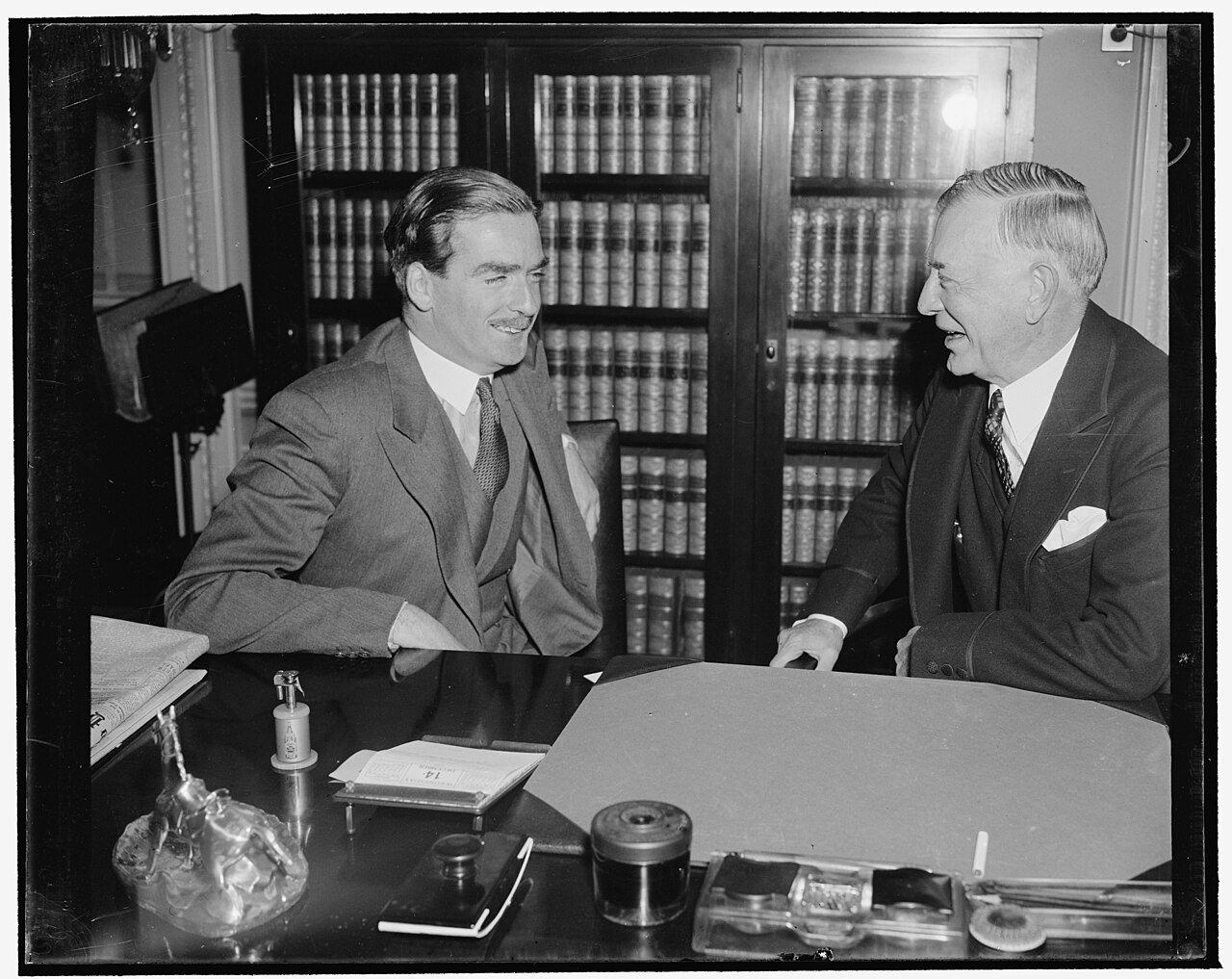 Two officials confer cordially across a desk before shelves of law books