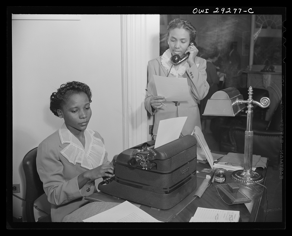 Two women in an office, one typing and one on the telephone reading a letter