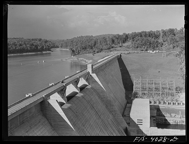 Norris Dam in Tennessee, a Tennessee Valley Authority public-works project. A massive concrete spillway curves down to a still reservoir, with the power house at right and forested hills beyond.