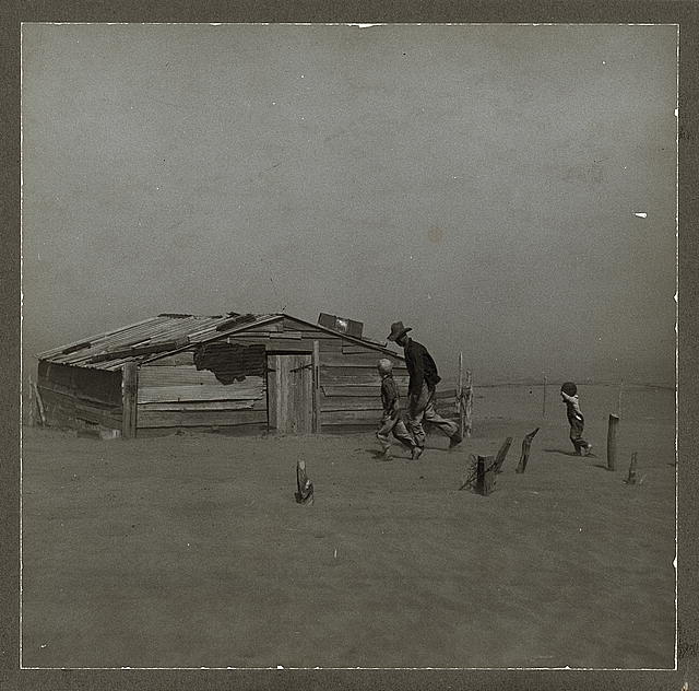 A father in a dark coat walks past a collapsed homestead with two young sons. Dust hangs in the air. Cimarron County, Oklahoma, April 1936.